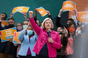 Andrea Horwath stands in front of a crown at a campaign rally. The crowd holds orange NDP signs and Ms. Horwath holds a microphone in one hand and her other fist upraised.